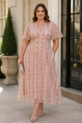 Front view of a blush pink plus size wedding guest midi dress with floral embroidered tulle, flutter sleeves, V-neckline and flowing midi skirt.