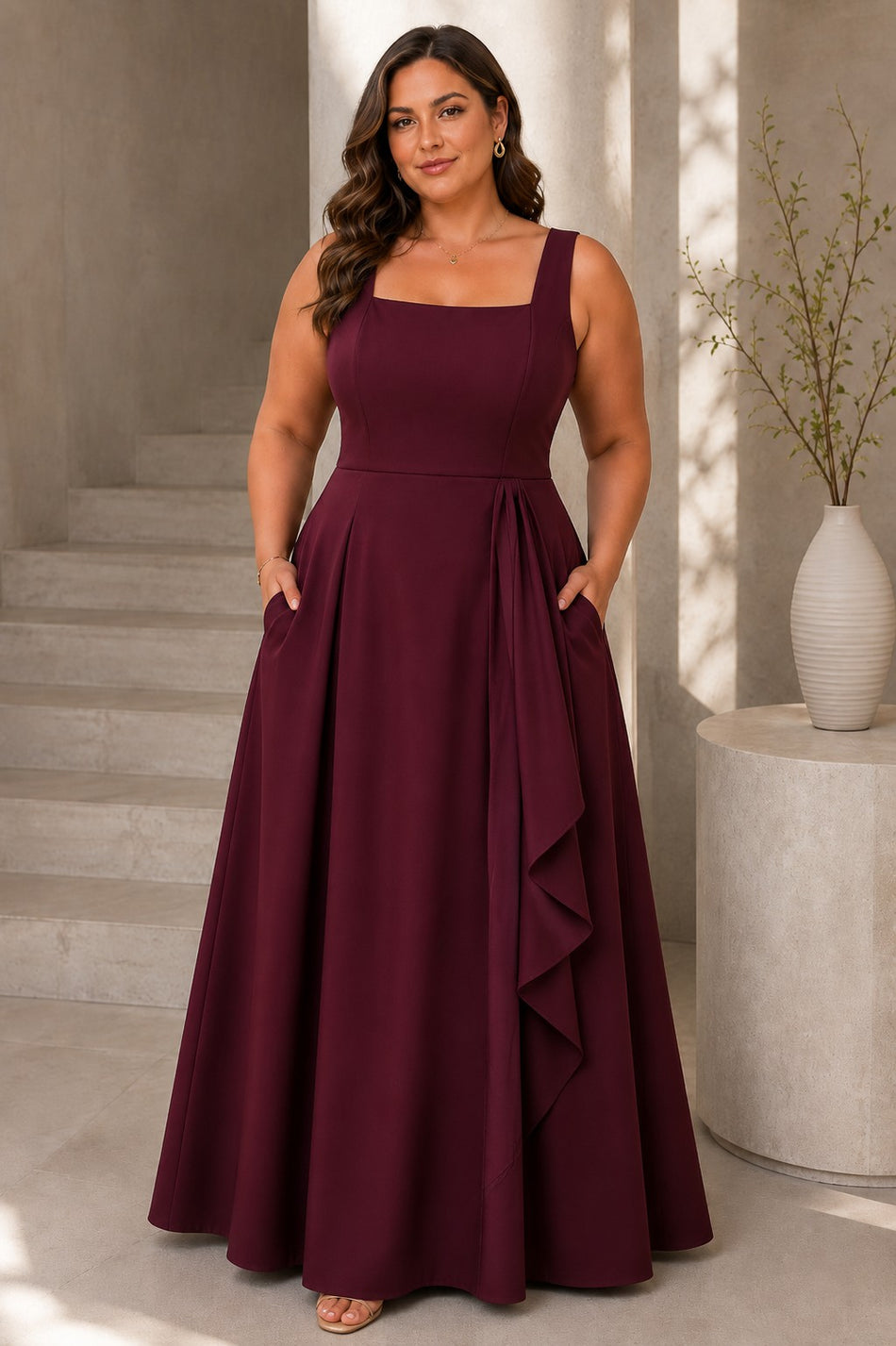 Plus size woman wearing a burgundy maxi formal dress with a square neckline, wide straps, full A-line skirt and cascading front drape.