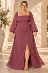 Plus size woman wearing a dusty rose pink evening maxi dress with square neckline, long sheer sleeves, draped waist and front split.